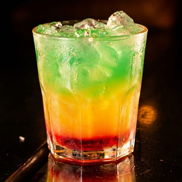 A vibrant layered cocktail with red, orange, and green colors, topped with ice, on a dark surface.