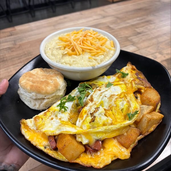 Black plate holding a cheesy breakfast omelet with potatoes, a biscuit, and a bowl of cheese-topped grits on a restaurant table.