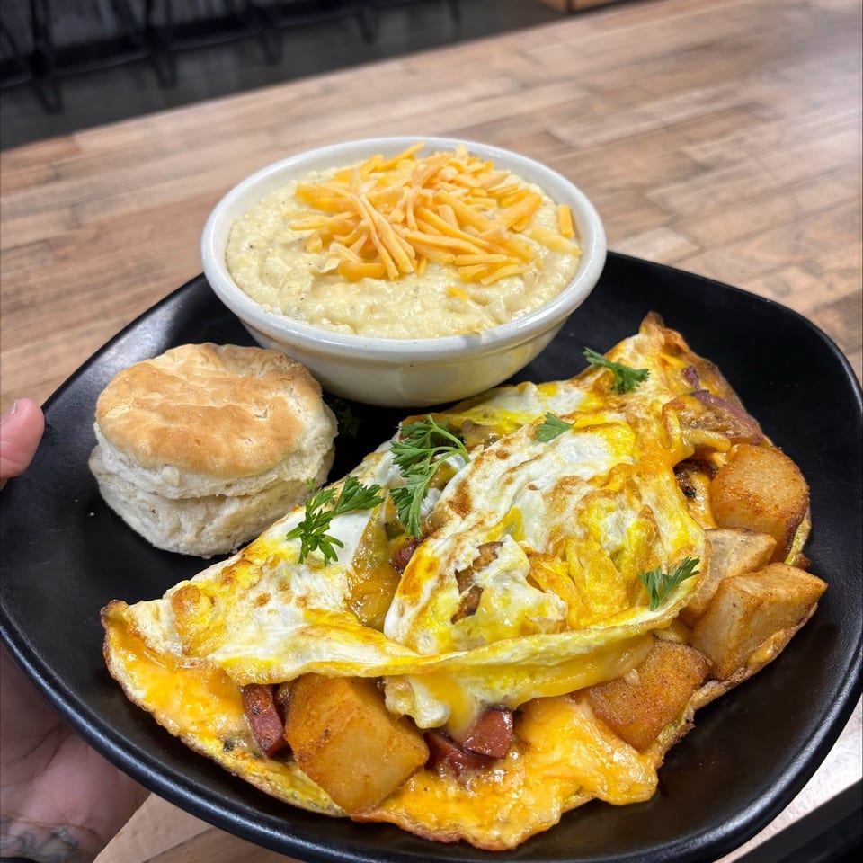 Black plate holding a cheesy breakfast omelet with potatoes, a biscuit, and a bowl of cheese-topped grits on a restaurant table.