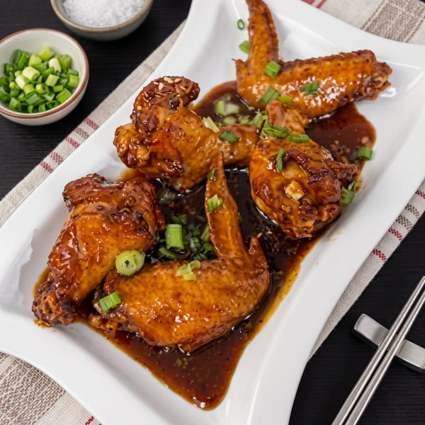 Glazed chicken wings garnished with green onions on a white platter, surrounded by bowls of salt, pepper, and chopped scallions.