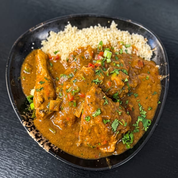 A black bowl of tender meat in rich orange curry sauce, garnished with herbs and scallions, served with rice.