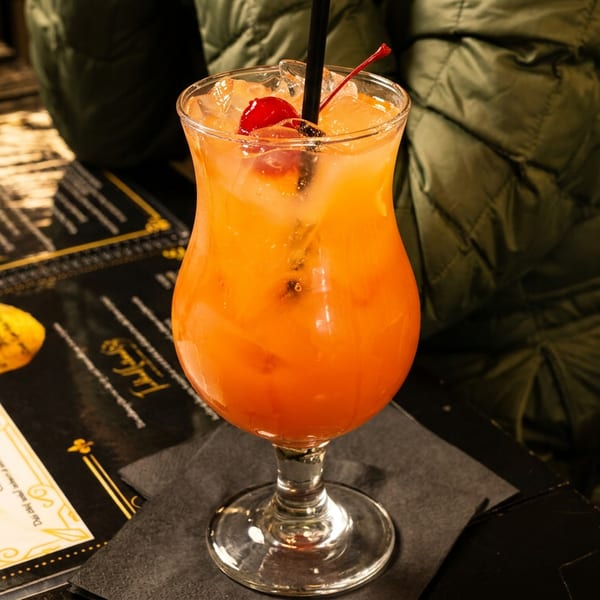 A vibrant orange cocktail with ice and cherries, next to menus on a dark table.