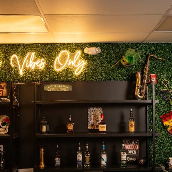 A bar area with a neon "Good Vibes Only" sign, bottles, and Mardi Gras decor on a green wall.